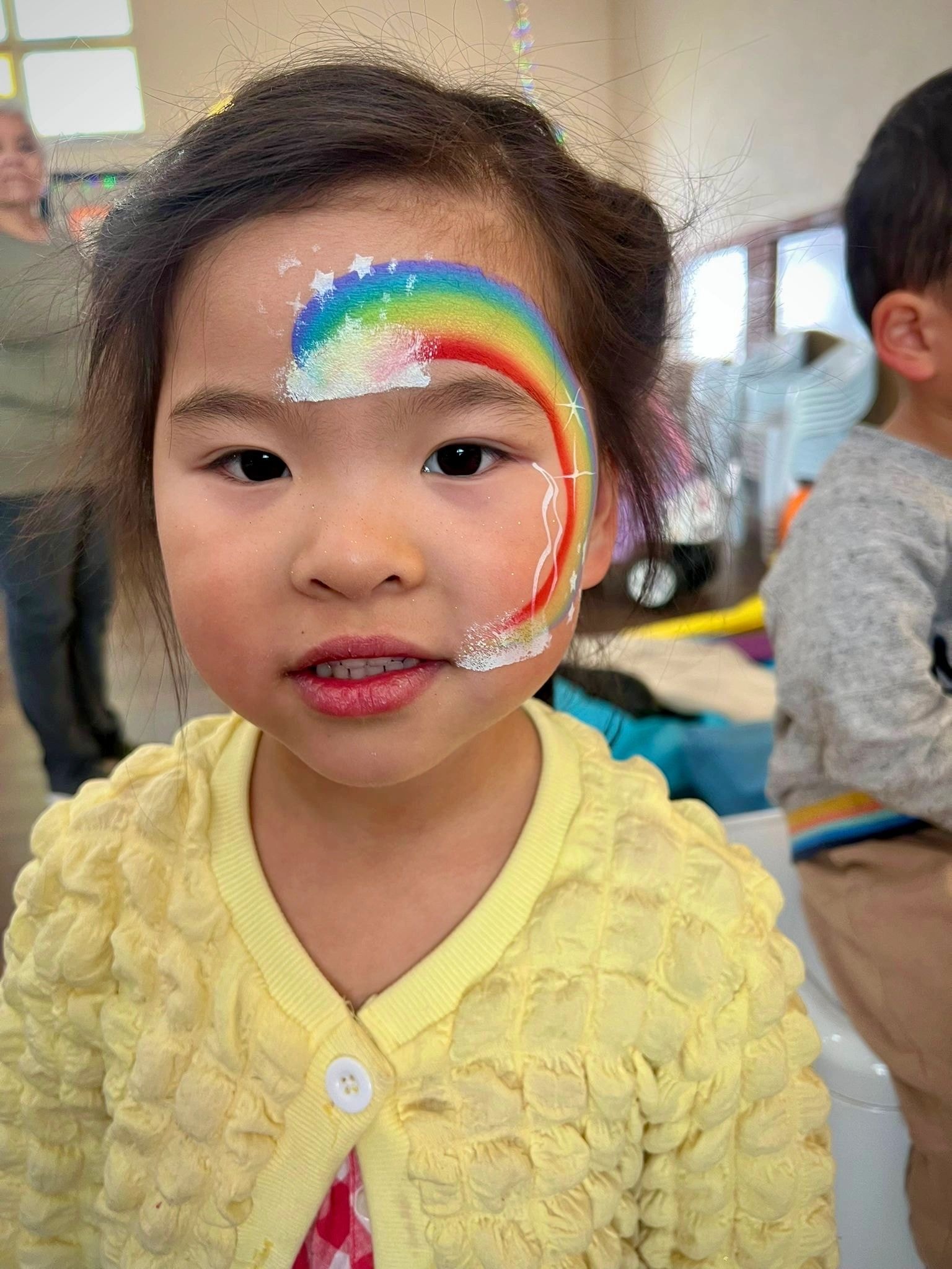 Child with rainbow face paint in yellow cardigan